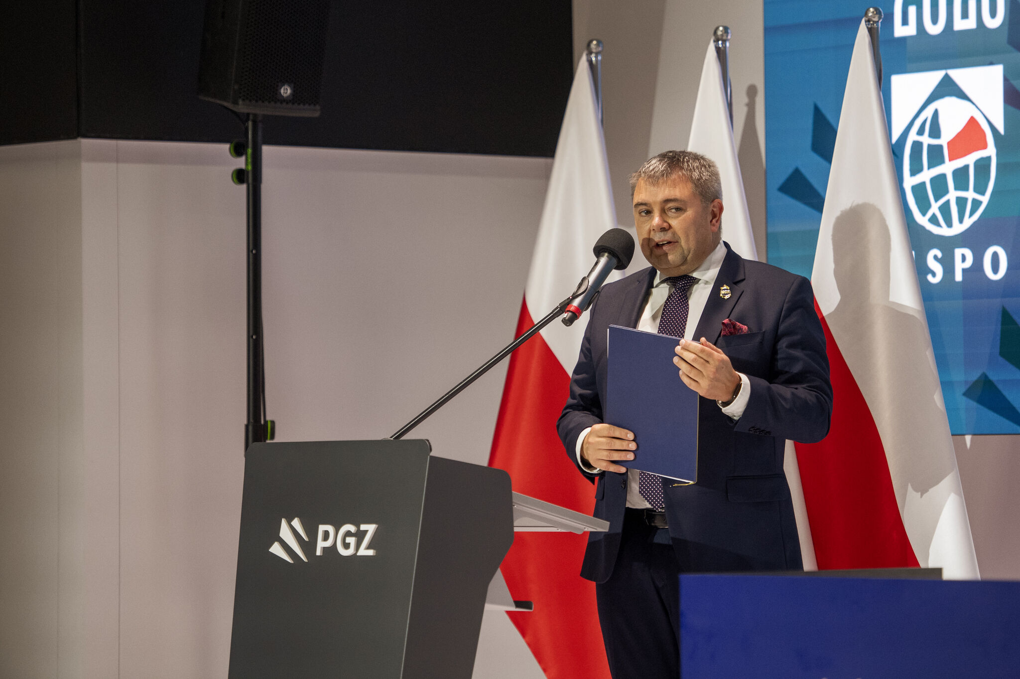 Przedstawiciel przy mównicy PGZ z teczką podczas podpisania listu intencyjnego — w tle polskie flagi i fragment logotypu MSPO