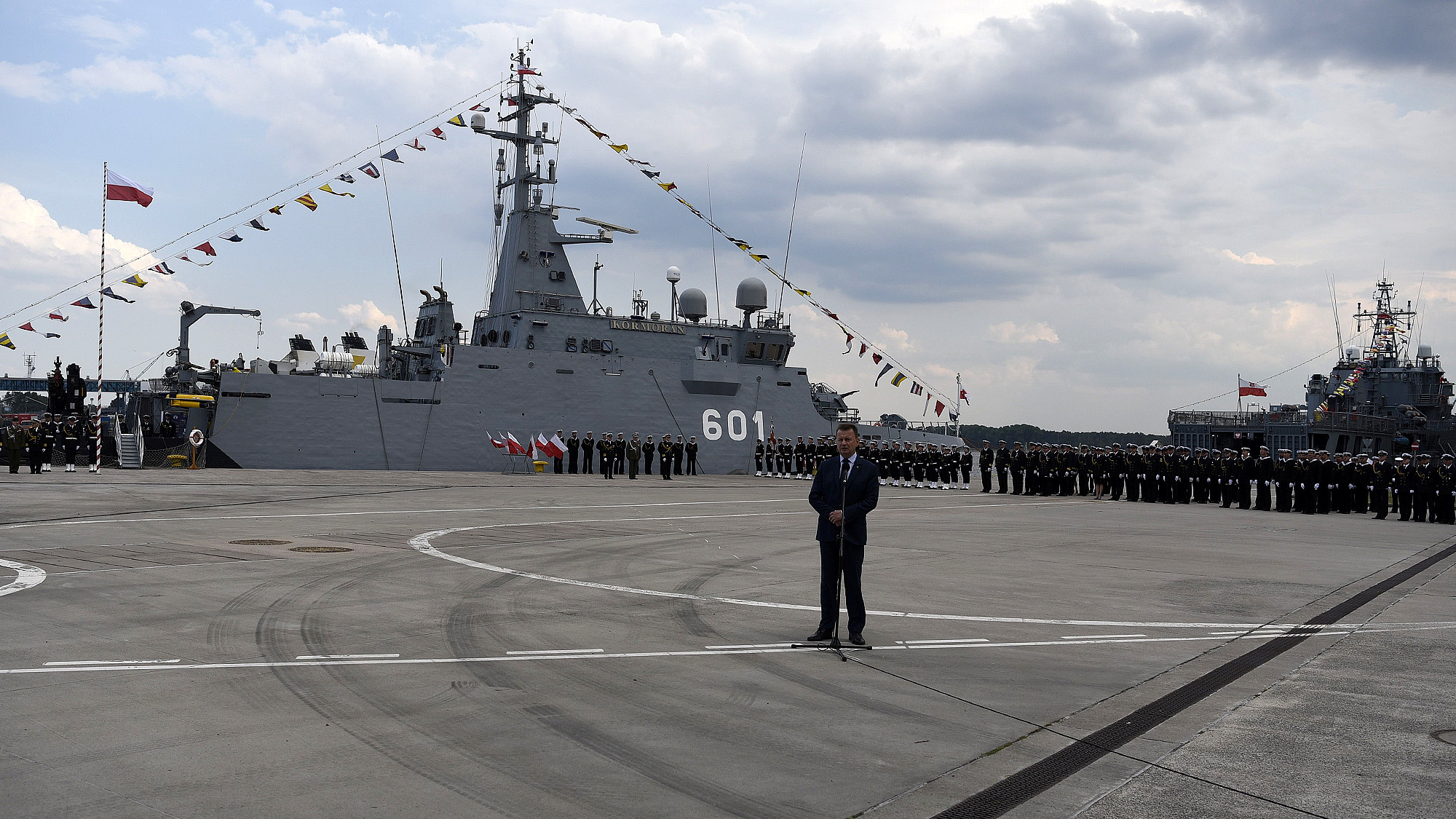 Przemówienie przy mikrofonie na nabrzeżu — w tle ORP Kormoran (601) udekorowany galą banderową