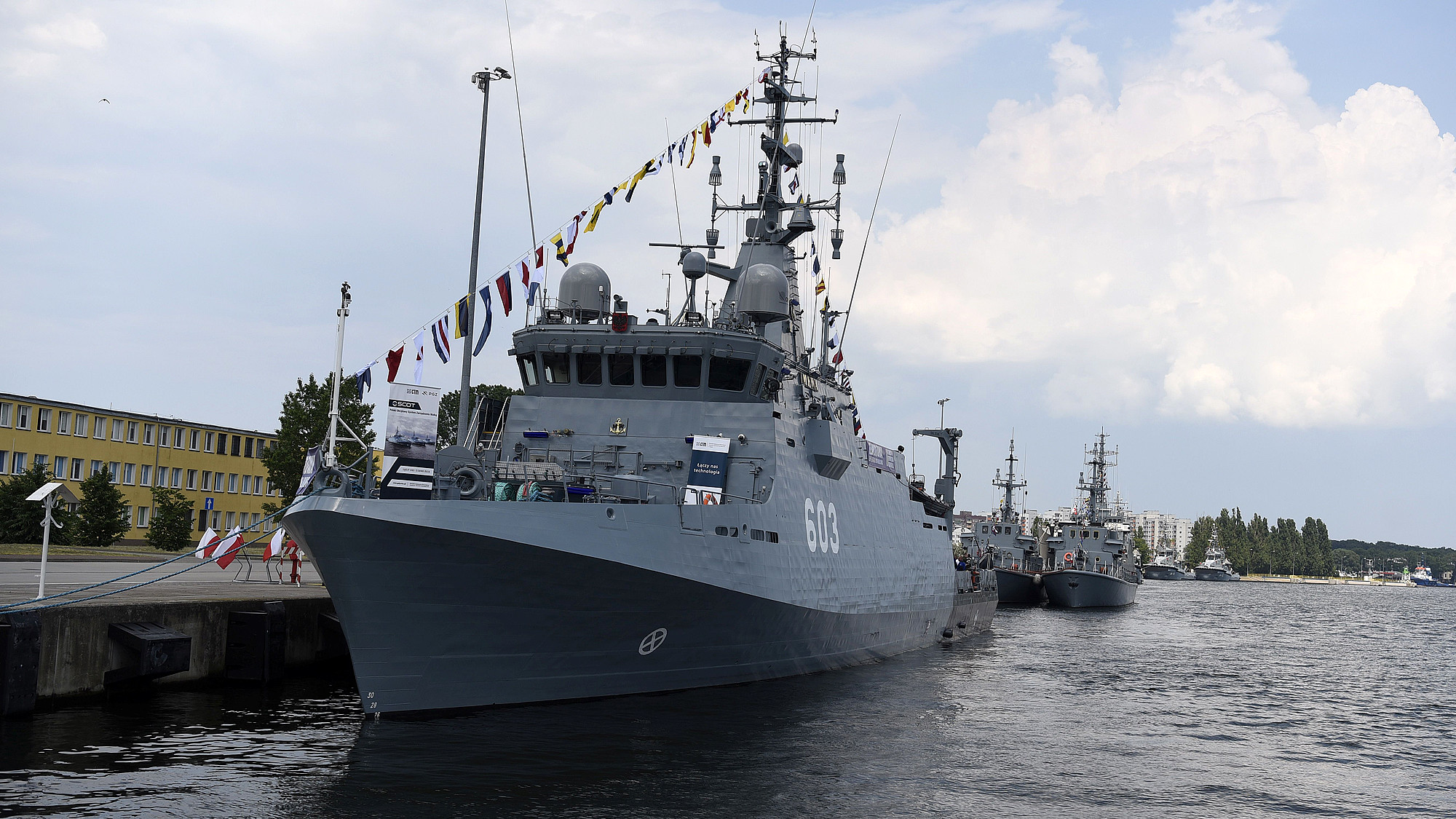 Niszczyciel min typu Kormoran II z galą banderową i banerami systemu SCOT, PGZ i CTM zacumowany przy nabrzeżu