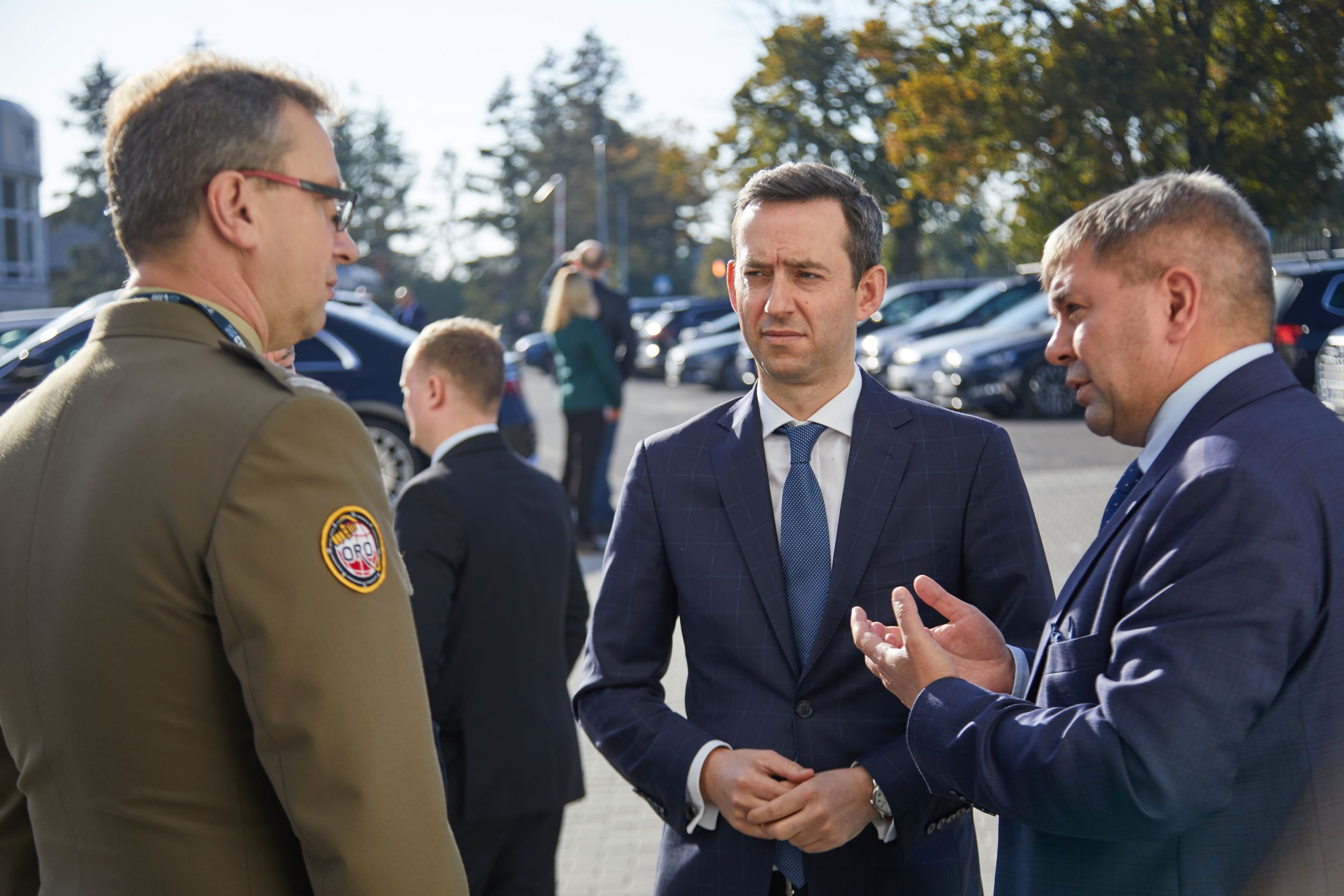 Oficer w mundurze wojskowym i dwaj mężczyźni w garniturach rozmawiający na parkingu podczas uroczystości otwarcia