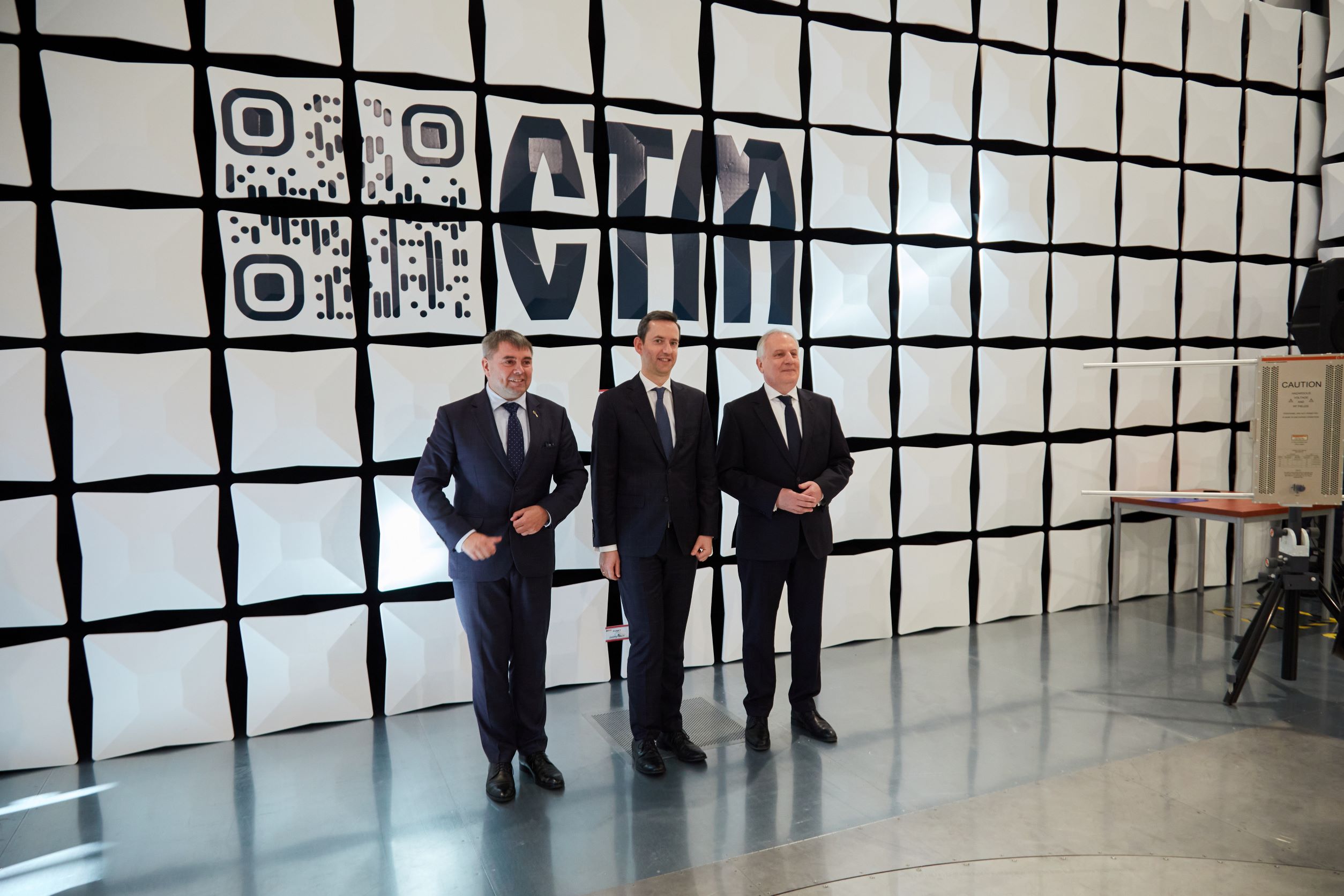 Trzech mężczyzn w garniturach w komorze bezechowej EMC LabNet na tle ściany z logo CTM i kodem QR