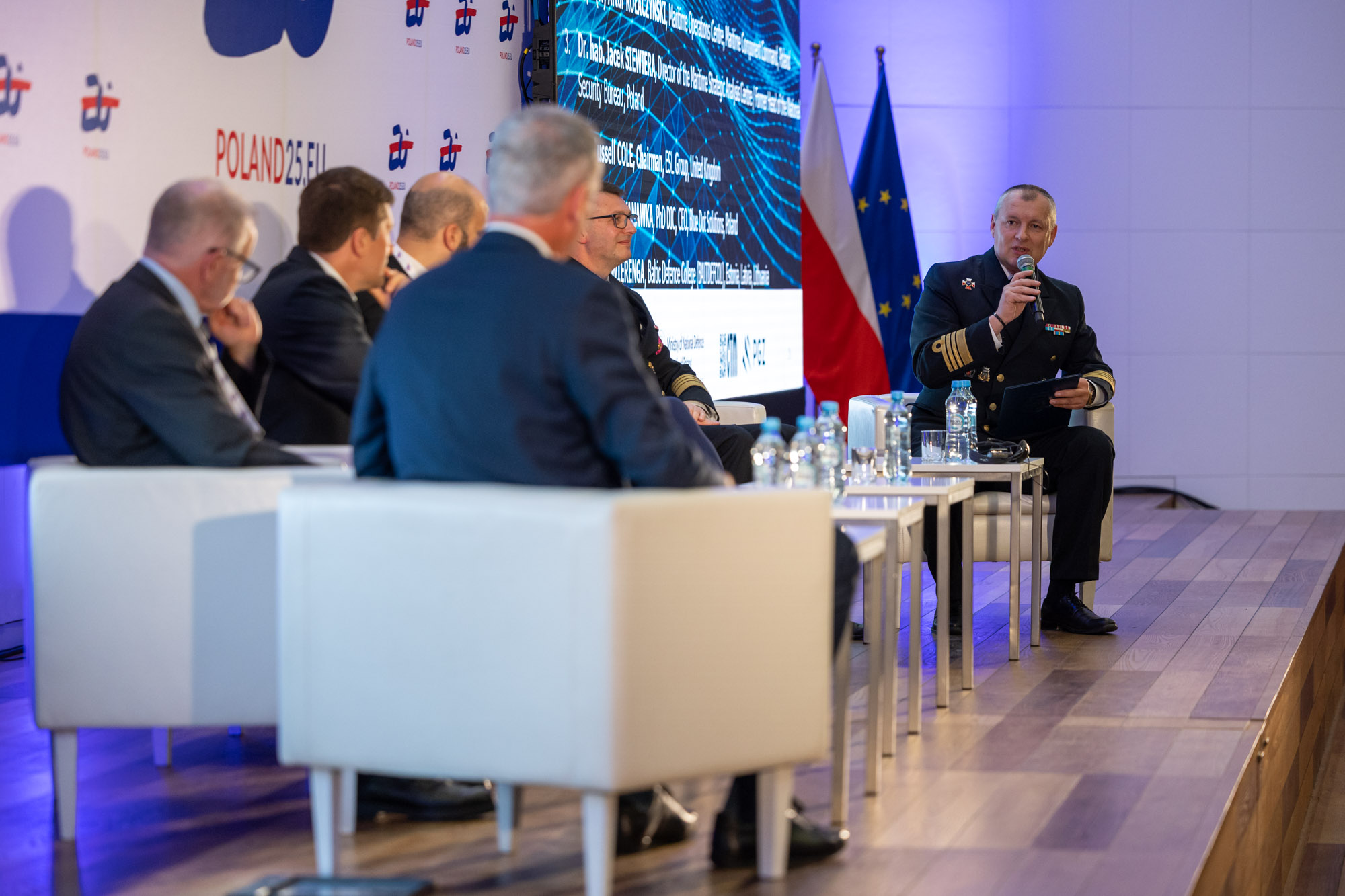 Oficer Marynarki Wojennej przemawia do mikrofonu na panelu — w tle flagi Polski i UE oraz ekran z logo CTM i PGZ