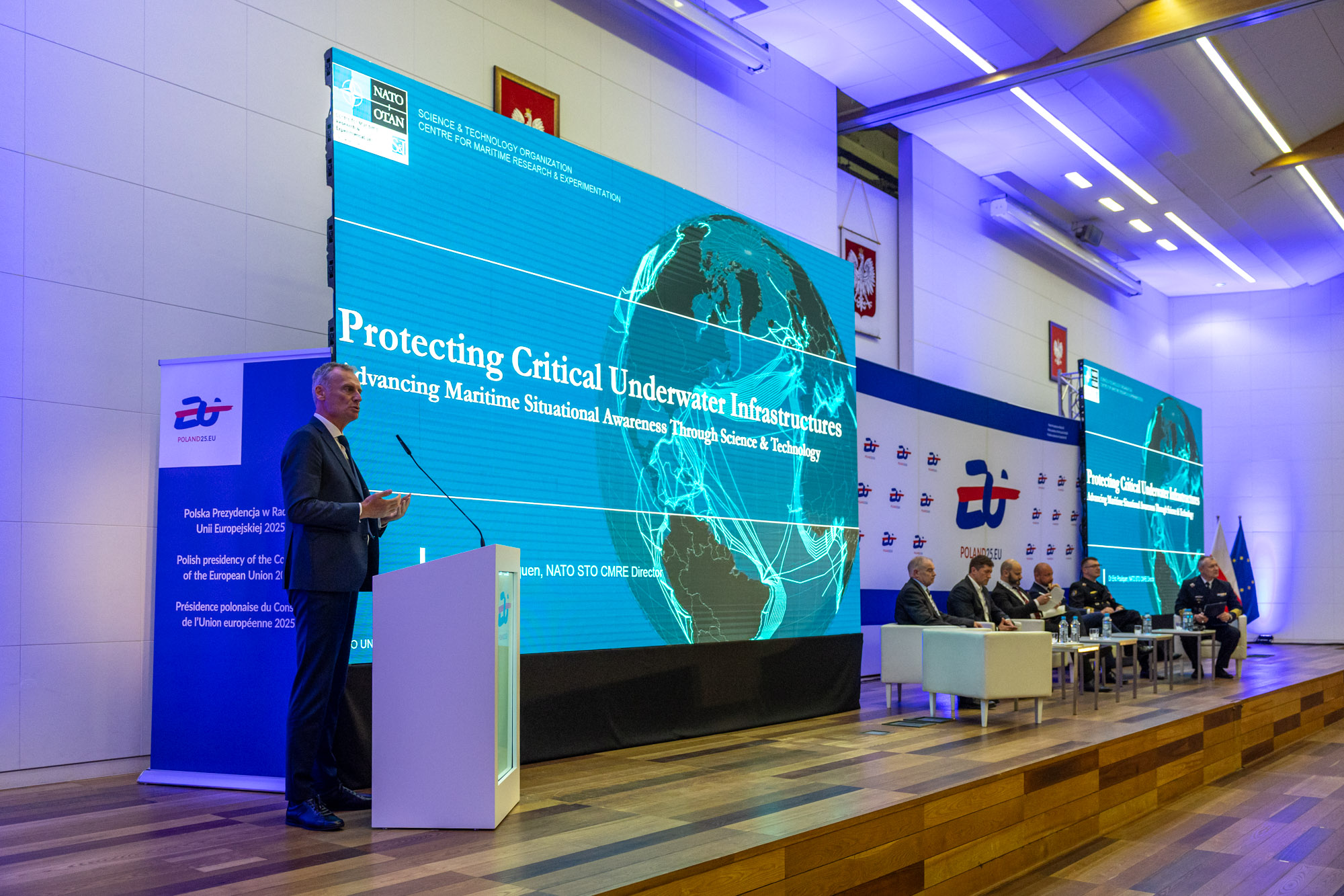 Prelegent przy mównicy przed ekranem NATO CMRE Protecting Critical Underwater Infrastructures — paneliści w fotelach po prawej