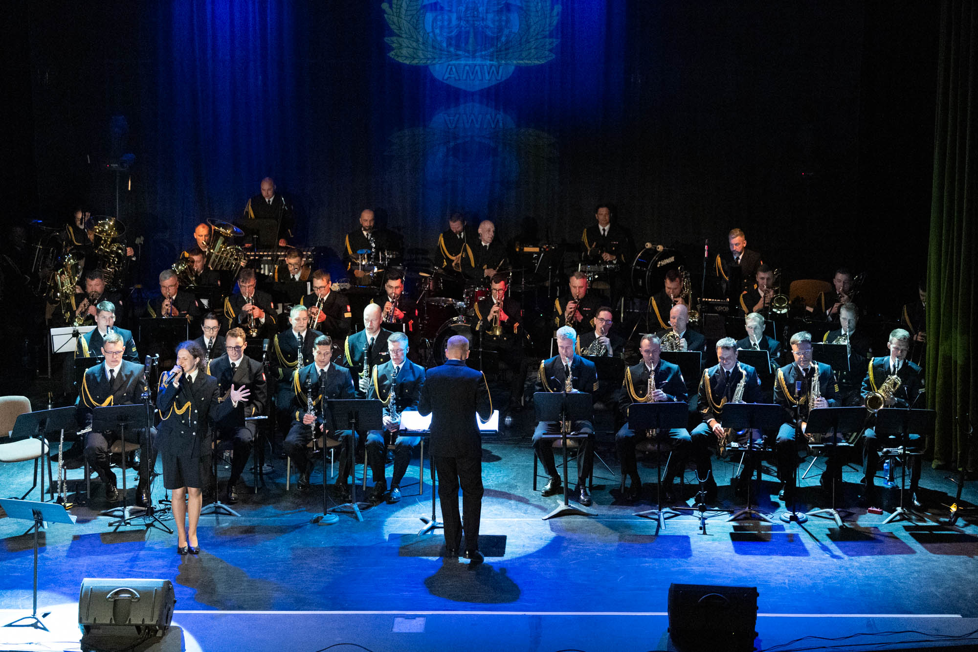 Orkiestra Reprezentacyjna Marynarki Wojennej na scenie z wokalistką i dyrygentem — niebieskie światło sceniczne i logo AMW