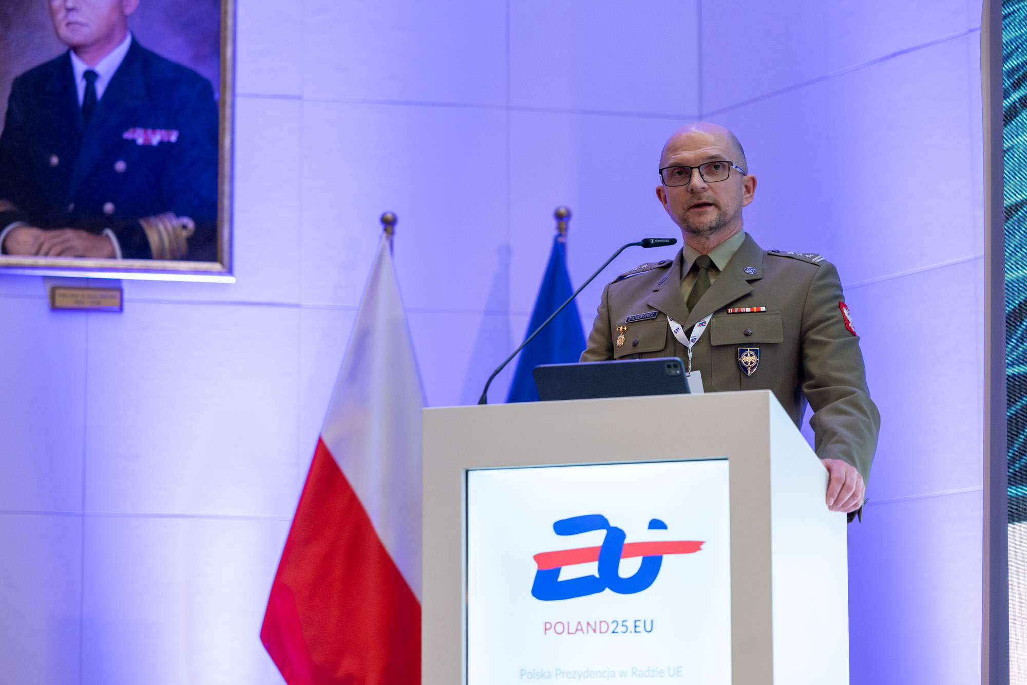 Pułkownik Wojska Polskiego przemawia przy mównicy z logo POLAND25.EU — w tle flagi Polski i portret oficera MW