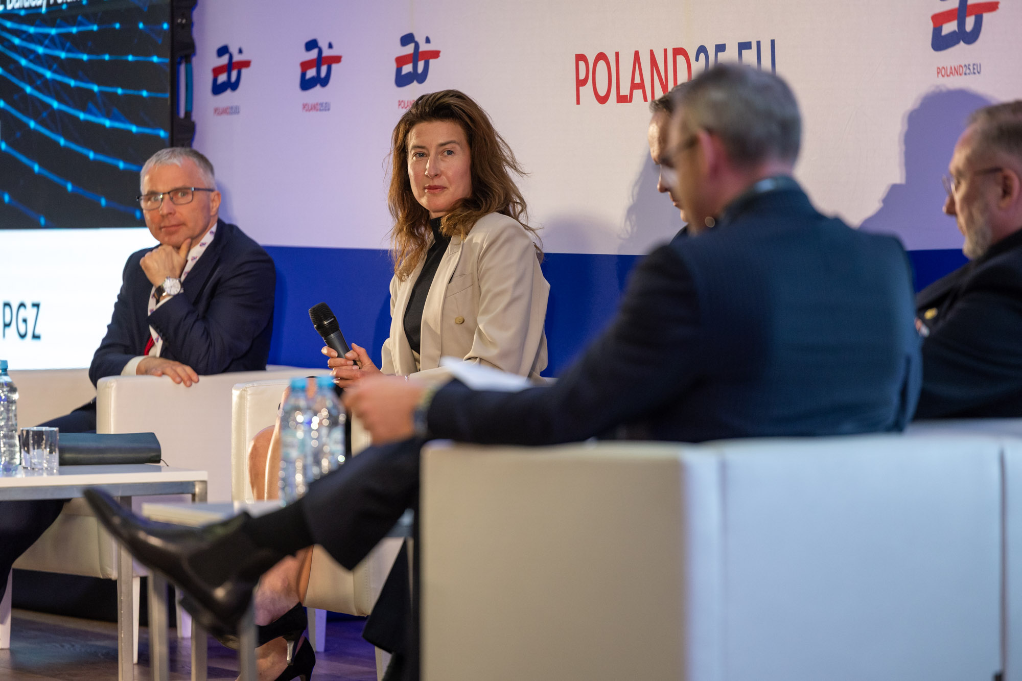 Panel dyskusyjny Digital Baltic — uczestniczka z mikrofonem w jasnej marynarce, w tle logo POLAND25.EU i PGZ na ekranie