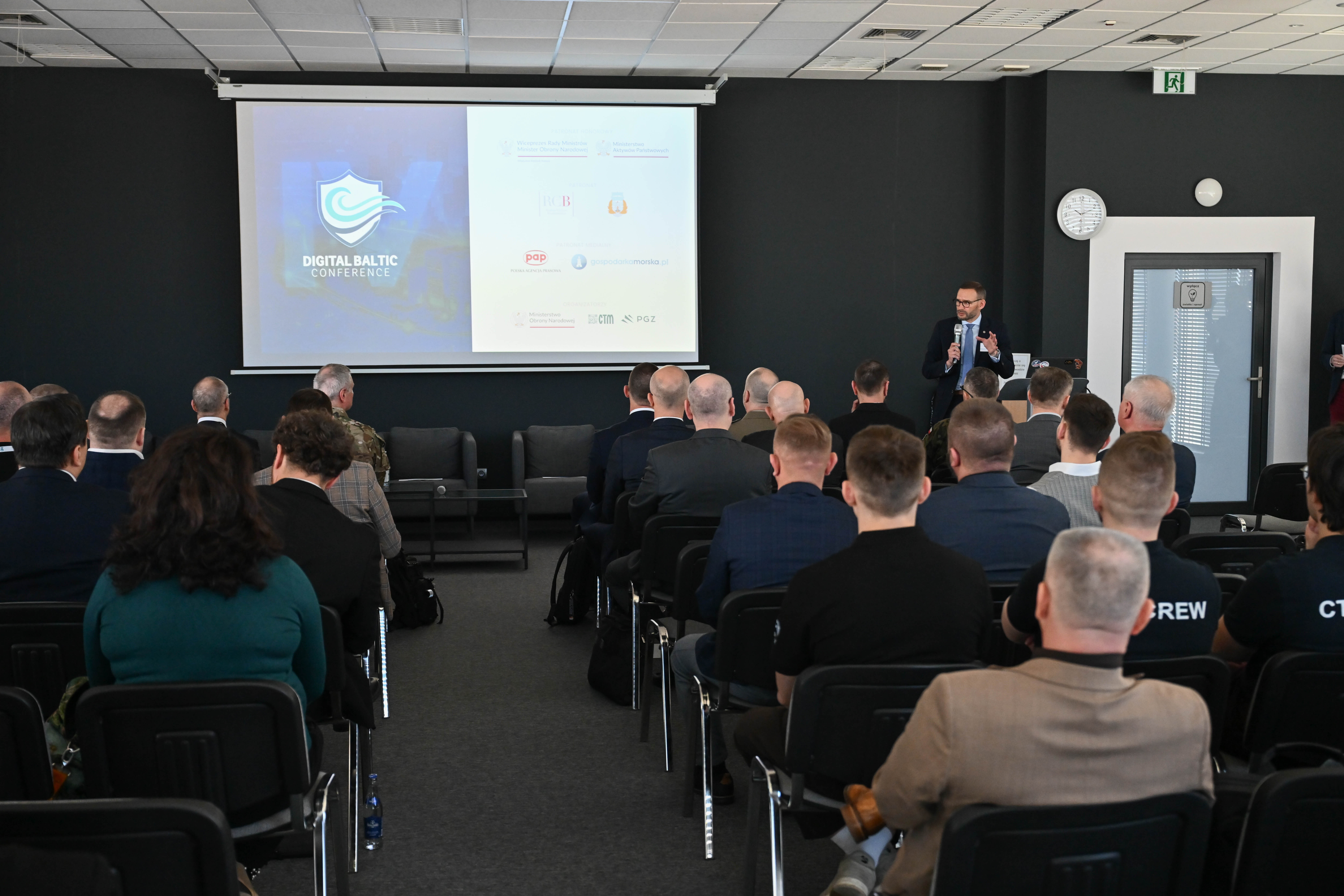 Prelegent przemawia do widowni Digital Baltic Conference — ekran z logo wydarzenia, uczestnicy w garniturach i mundurach