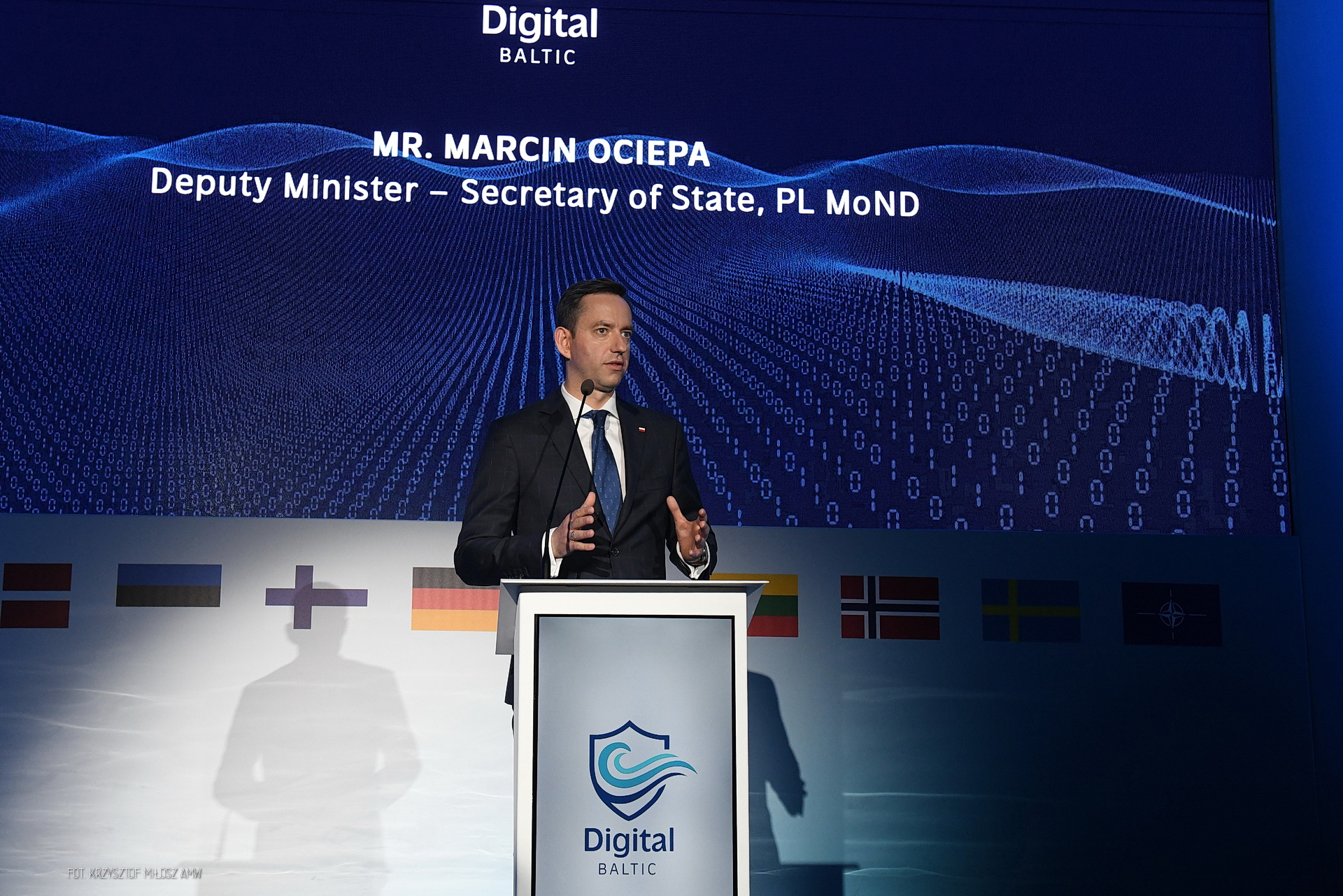 Marcin Ociepa przemawia przy mównicy Digital Baltic na tle ekranu i flag państw partnerskich oraz NATO