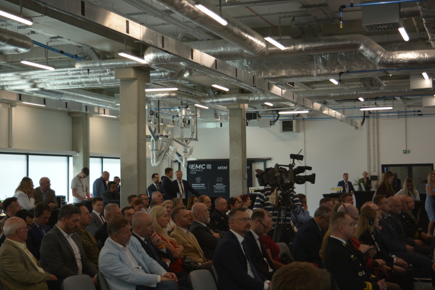 Publiczność uroczystości otwarcia CEM w nowoczesnej hali technologicznej — goście w fotelach, w tle baner EMC Defence Hub