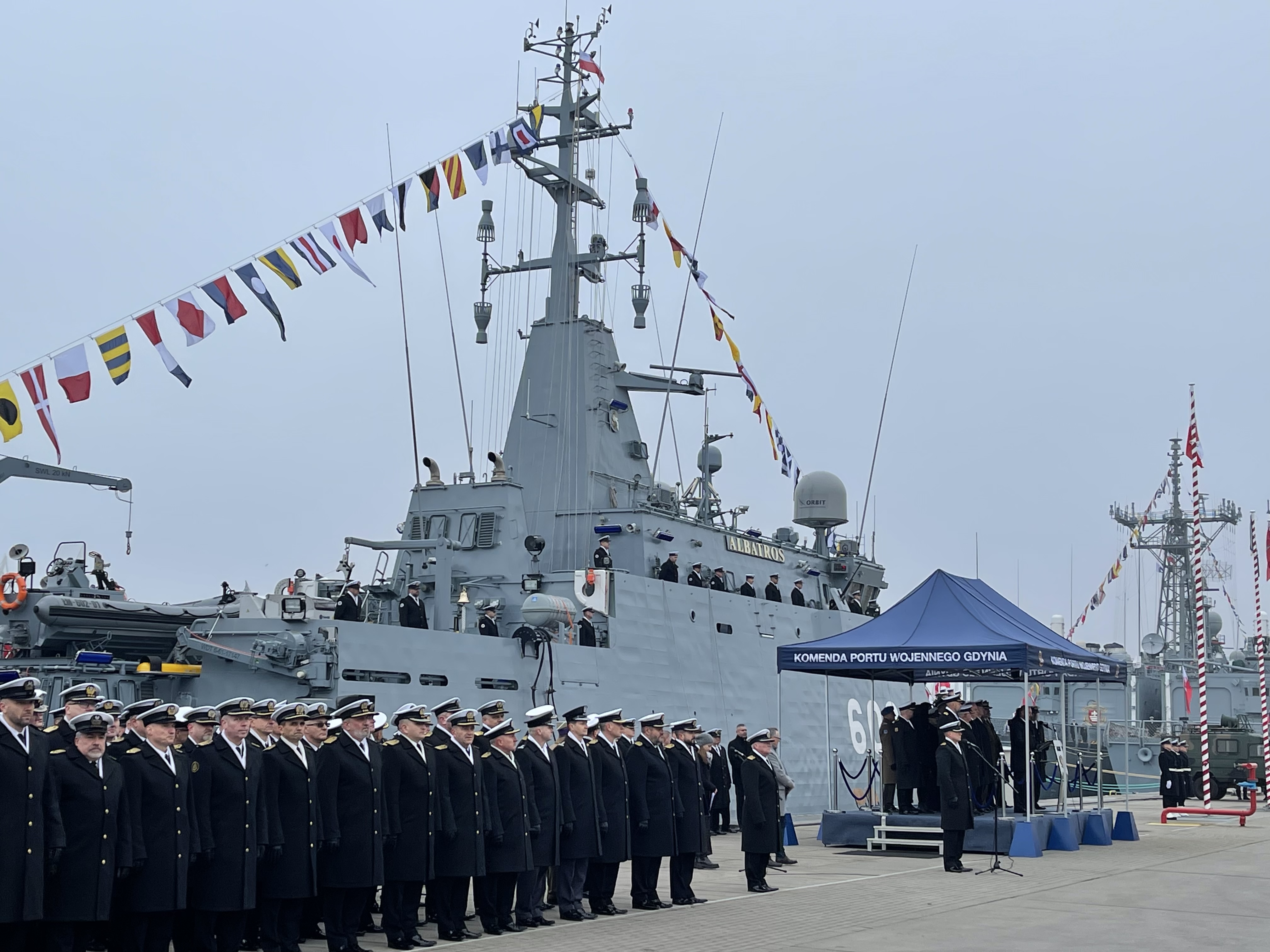 Oficerowie Marynarki Wojennej w szyku na nabrzeżu podczas uroczystości — ORP Albatros (602) z galą banderową i namiot KPW Gdynia
