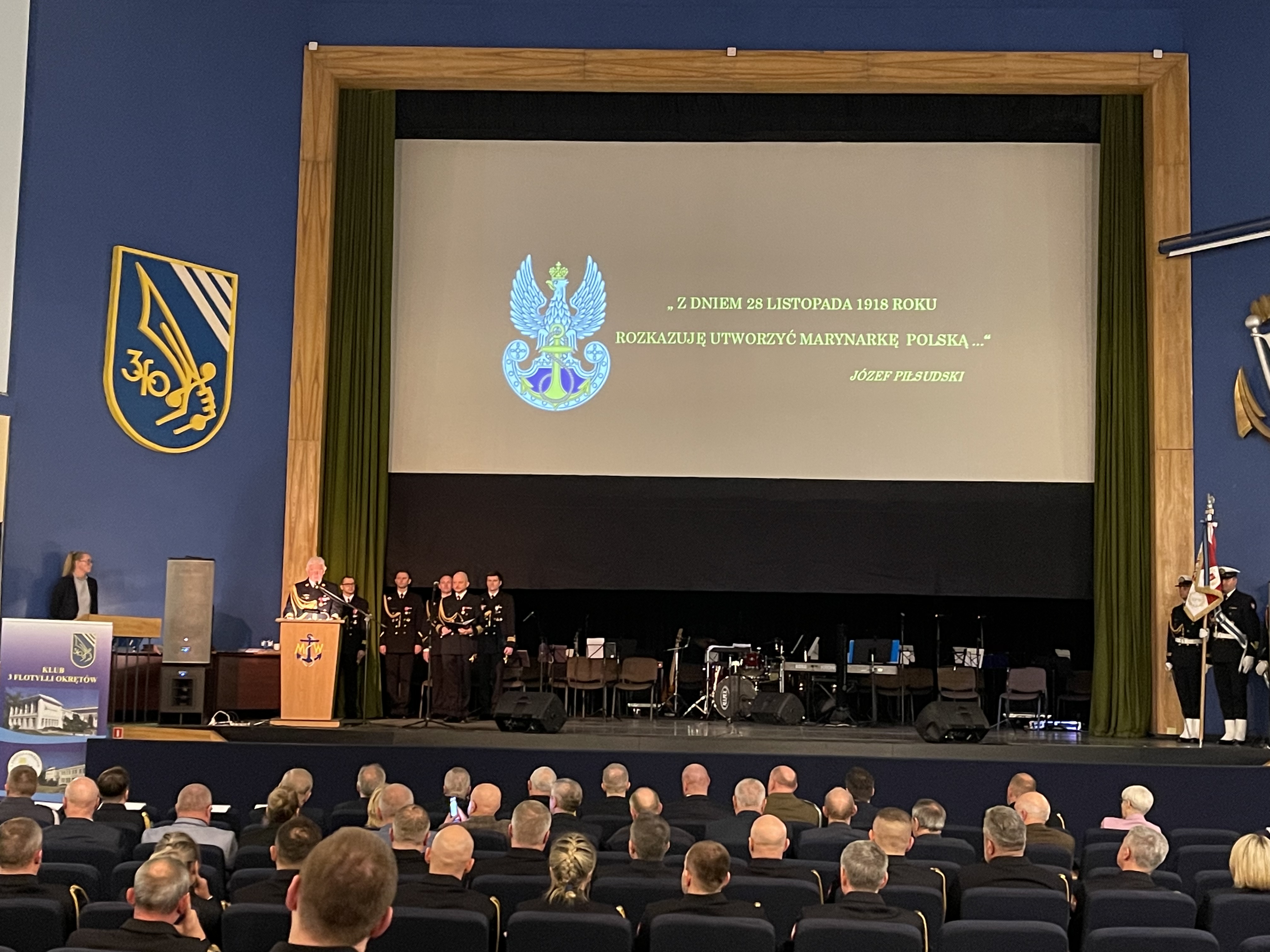 Ceremonia w Klubie 3 Flotylli Okrętów — oficer przemawia przy pulpicie, ekran z godłem Marynarki Wojennej i cytatem Józefa Piłsudskiego