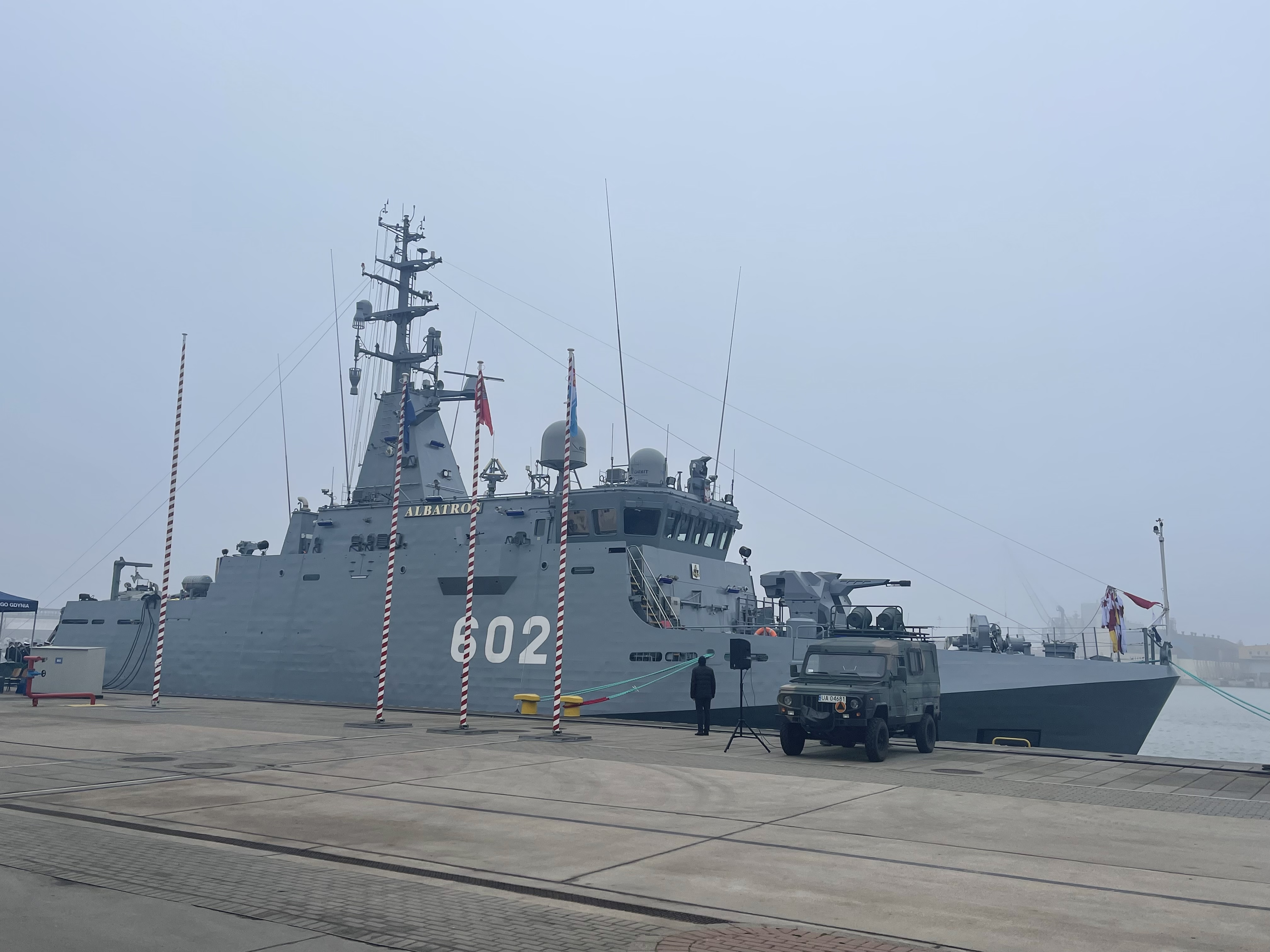 ORP Albatros (602) zacumowany przy nabrzeżu w mglisty dzień — wojskowy pojazd terenowy i maszty na pierwszym planie