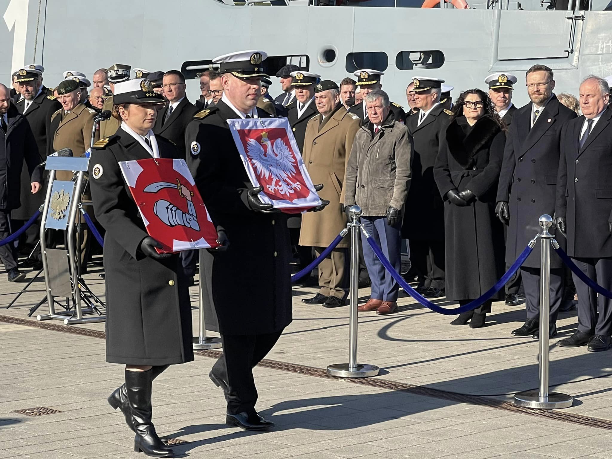 Oficerowie Marynarki Wojennej niosą uroczyście proporzec i banderę podczas ceremonii na nabrzeżu