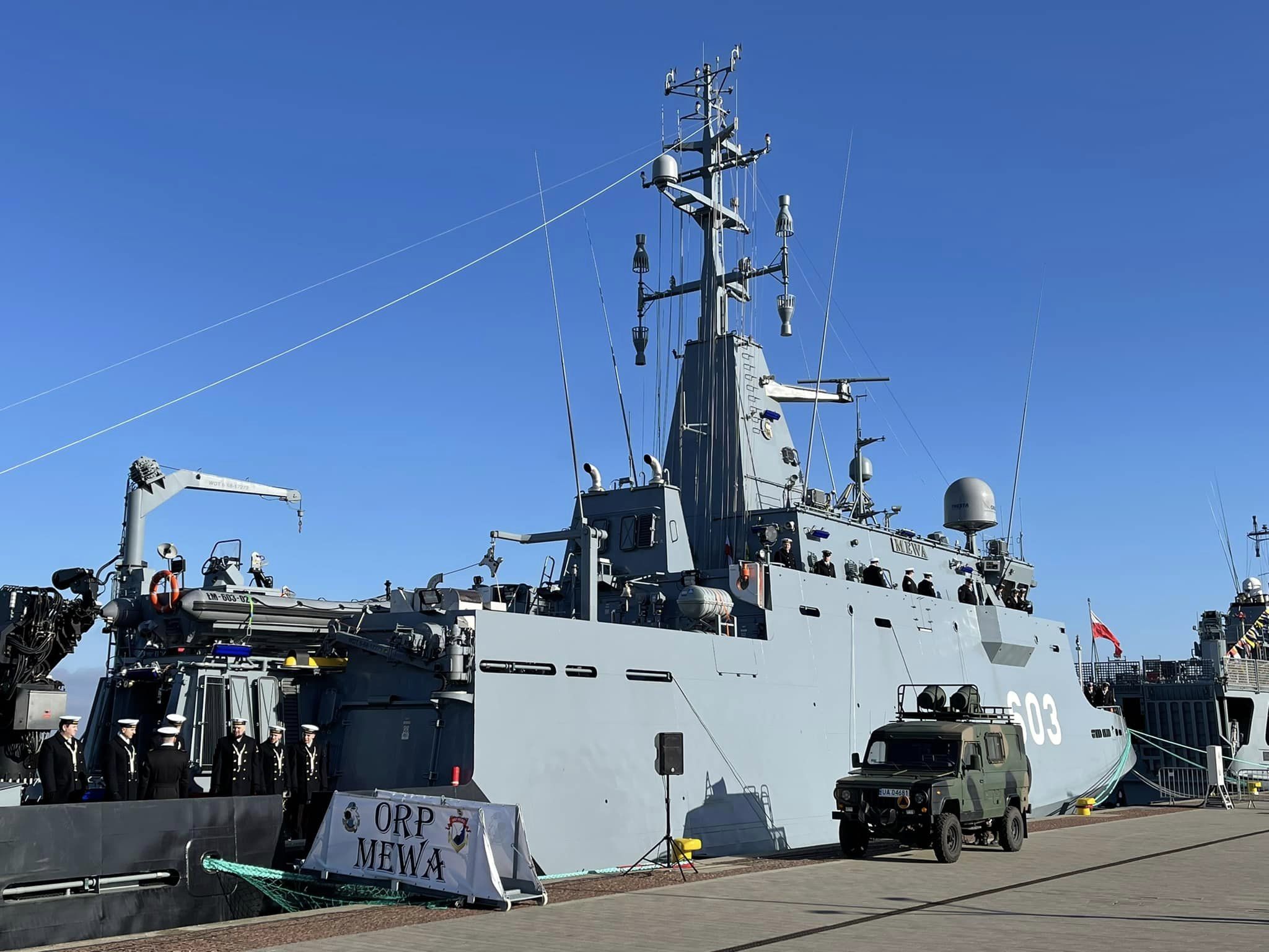 ORP Mewa (603) podczas uroczystości — tablica z nazwą okrętu na nabrzeżu, marynarze na pokładzie i pojazd wojskowy