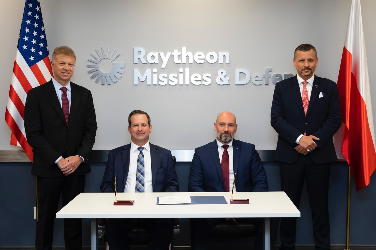 Czterej przedstawiciele PGZ i Raytheon przy stole z dokumentami na tle logo Raytheon Missiles & Defense i flag Polski oraz USA