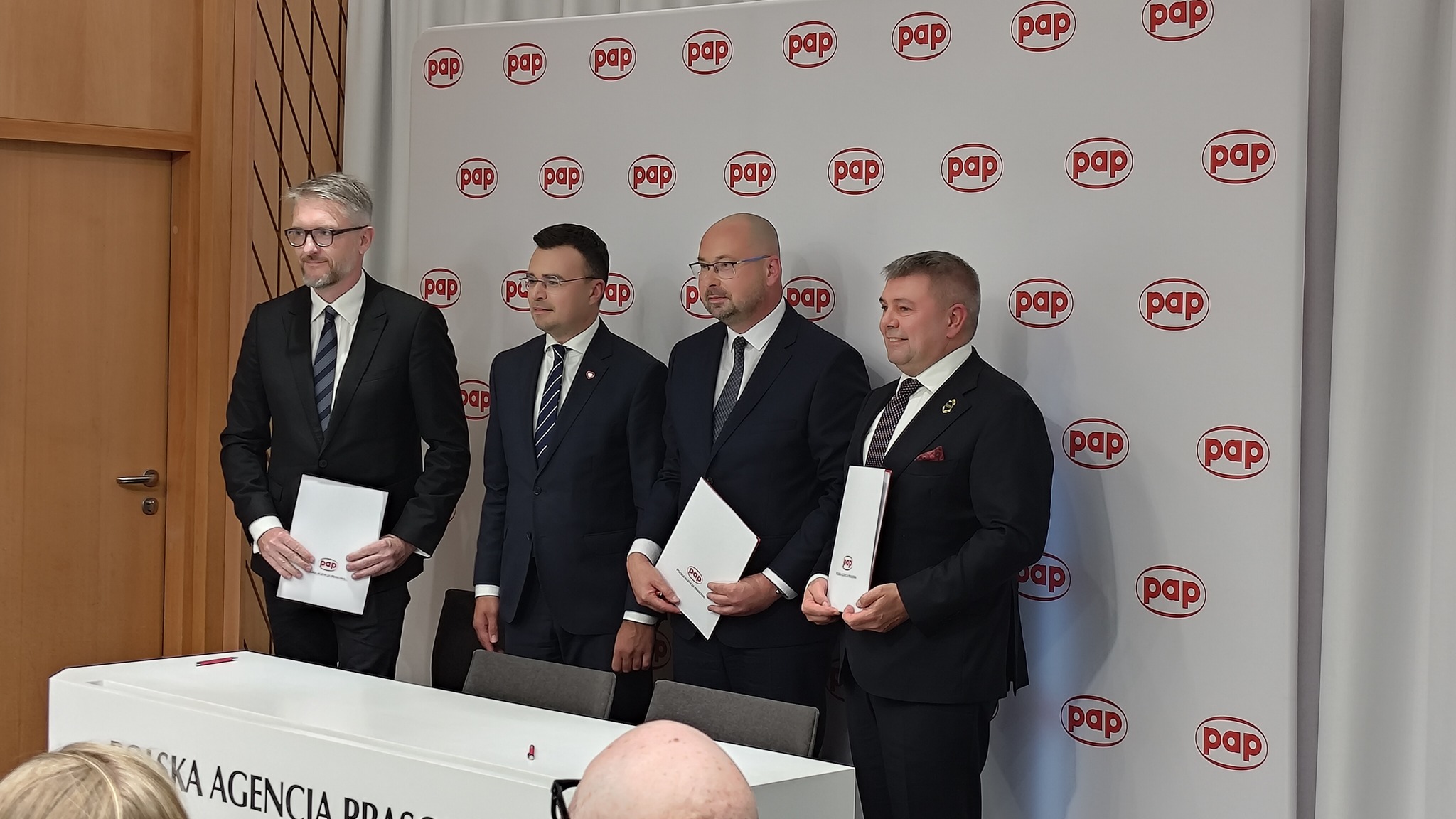 Czterech mężczyzn w garniturach z dokumentami za stołem Polskiej Agencji Prasowej na tle ścianki PAP