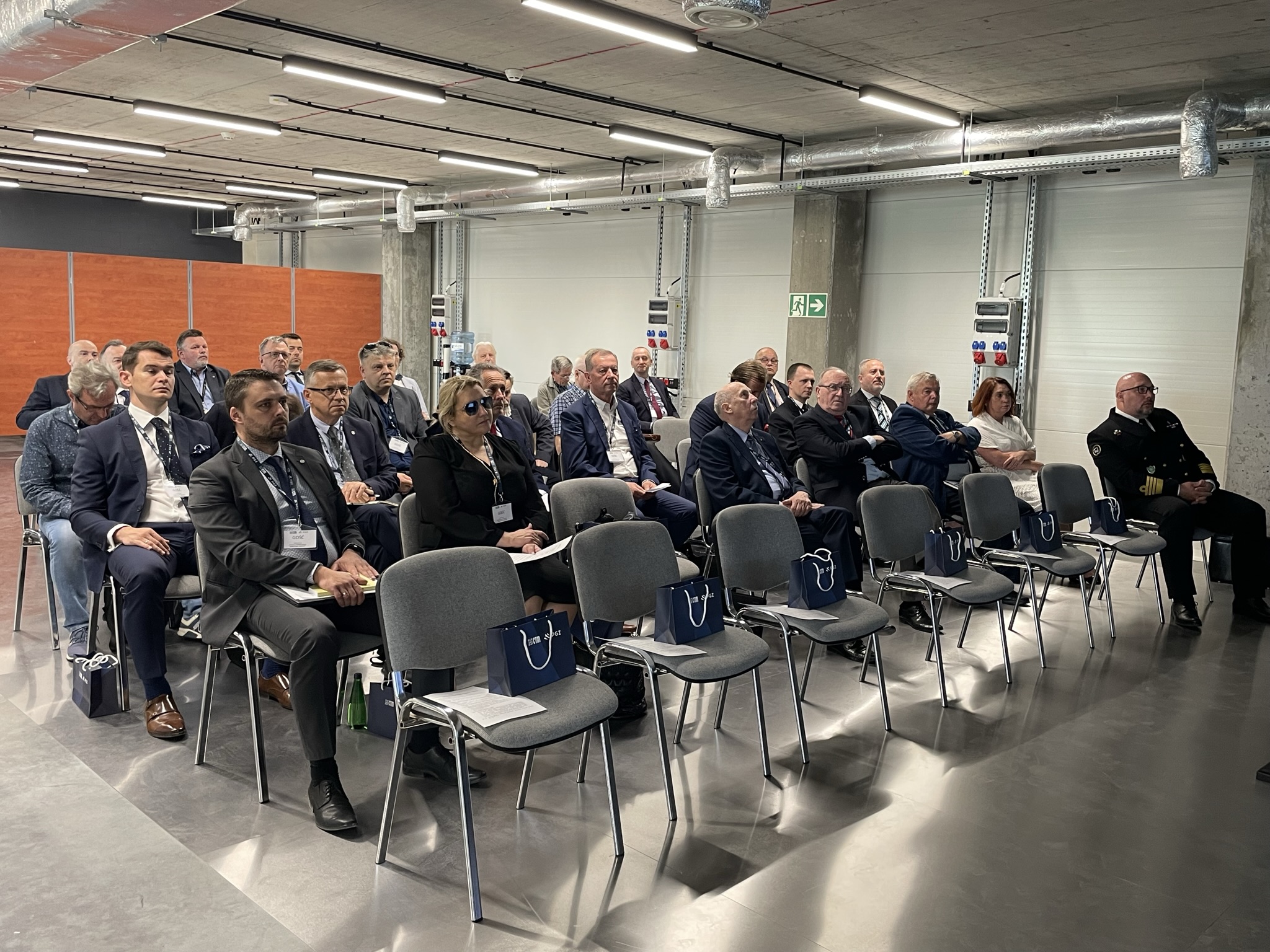 Uczestnicy konferencji o programie Miecznik w sali z betonowymi ścianami — widownia w garniturach i mundurze marynarskim