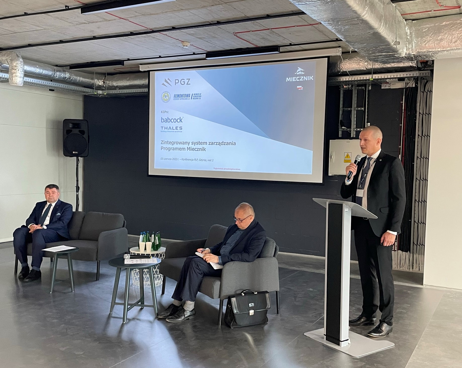 Wystąpienie przy ekranie z tytułem Zintegrowany system zarządzania Programem Miecznik i logotypami PGZ, Remontowa Shipbuilding, Babcock i Thales