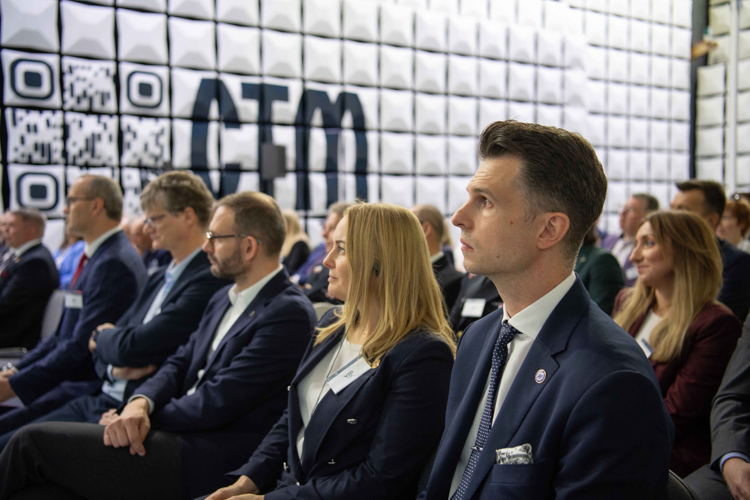 Widownia uroczystości w komorze bezechowej CTM — goście w garniturach na tle ściany z logo CTM i kodem QR