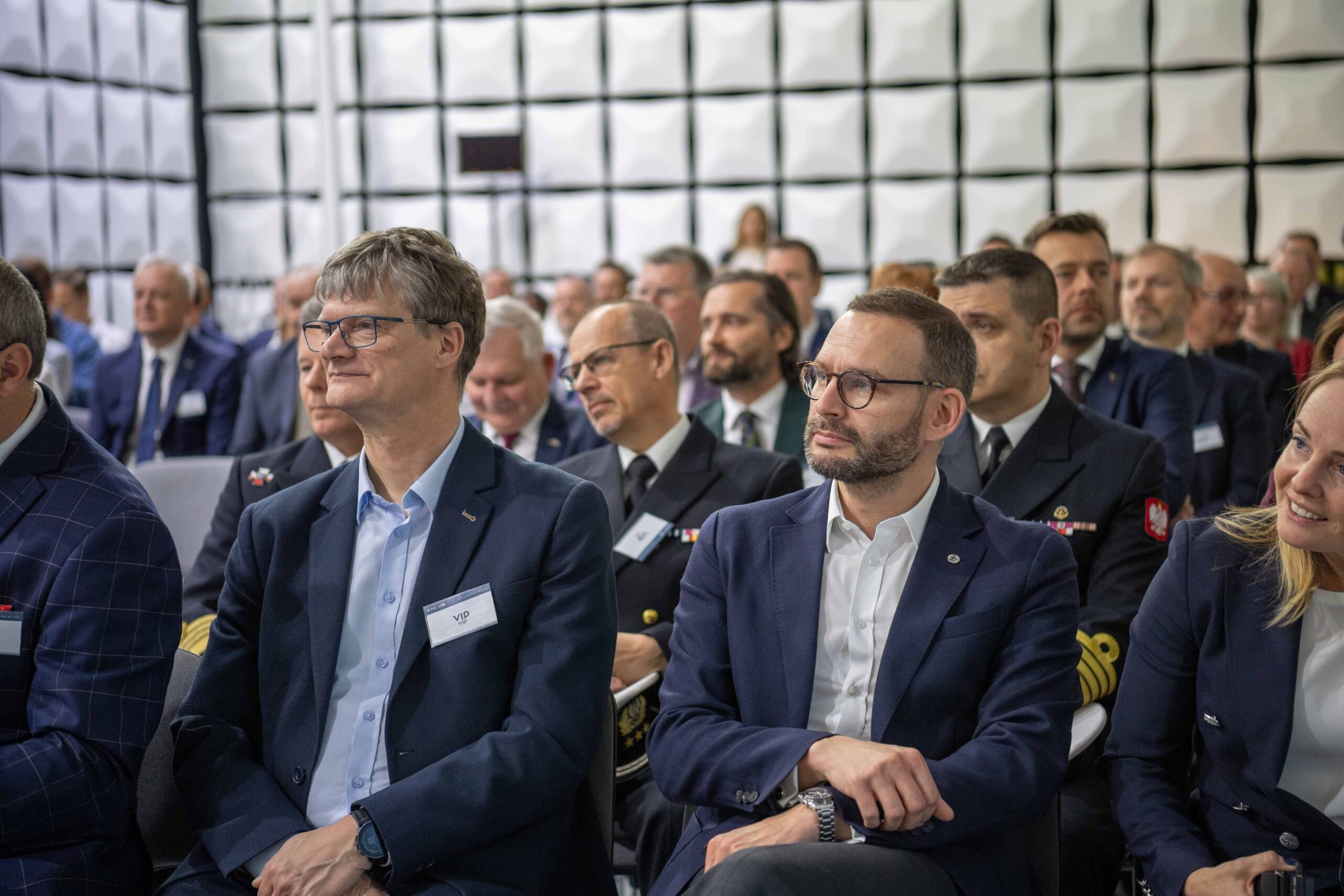 Uczestnicy uroczystości w komorze bezechowej — cywile w garniturach i oficerowie Marynarki Wojennej na tle paneli akustycznych