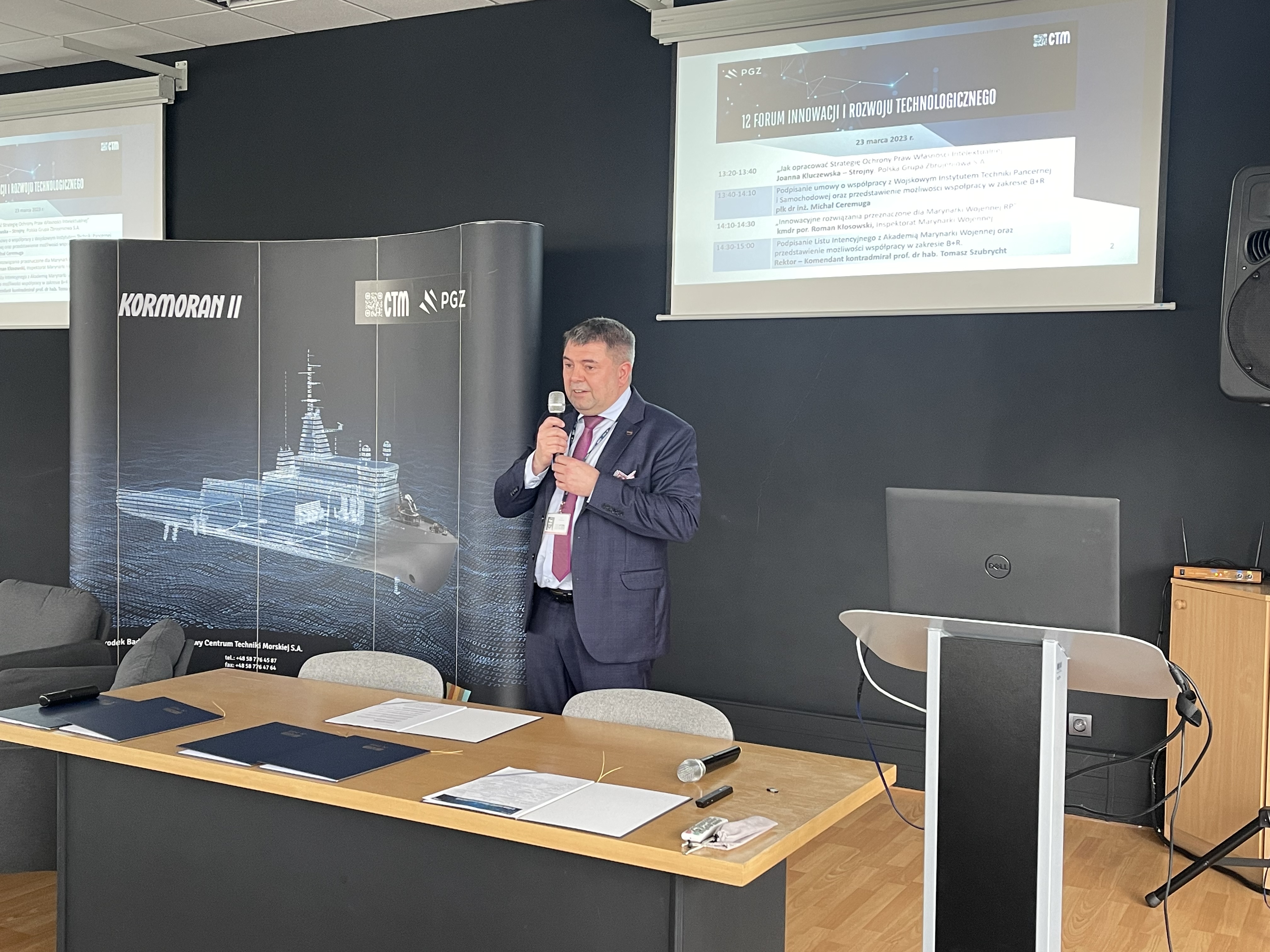 Prelegent przy mównicy podczas XII Forum Innowacji — baner Kormoran II i ekran z agendą wydarzenia